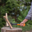 Persona che spacca legna con un'ascia Fiskars X7 su un ceppo di legno in un ambiente esterno, indossando una camicia grigia e jeans. Dettaglio dell'azione di spaccatura della legna con schegge che volano.